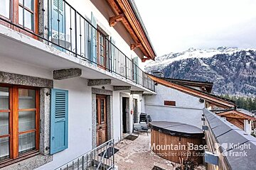 A charming mountain chalet with a balcony, hot tub, and blue shutters, set against a backdrop of snow-capped mountains.