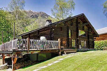 A rustic wooden cabin with a large deck sits on a grassy hill, surrounded by trees with a snow-capped mountain in the background.