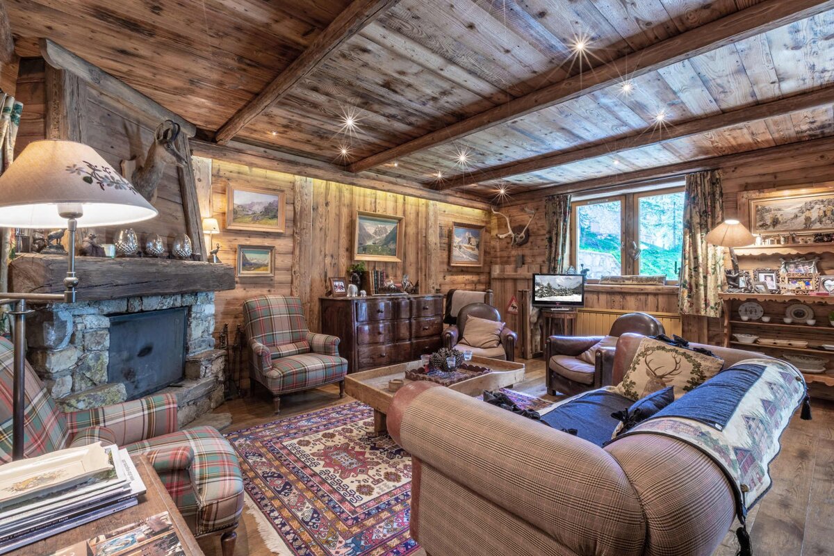 Cozy, rustic wood-paneled lodge living room with a stone fireplace, plaid chairs, leather sofas, and an intricate patterned rug.