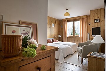 A cozy, wood-paneled bedroom with two single beds, a window with curtains, an armchair, and a TV. A dresser with art and flowers is in the foreground.