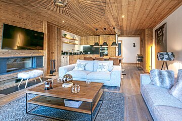 A modern open-plan living room and kitchen with wood-paneled walls, ceiling, and floor, featuring gray sofas, a fireplace, and a dining area.