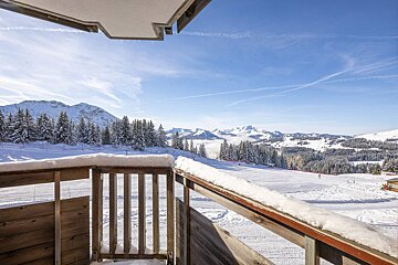 A snow-covered balcony overlooks a sunny mountain ski resort with pine forests, majestic peaks, and a bright blue sky.