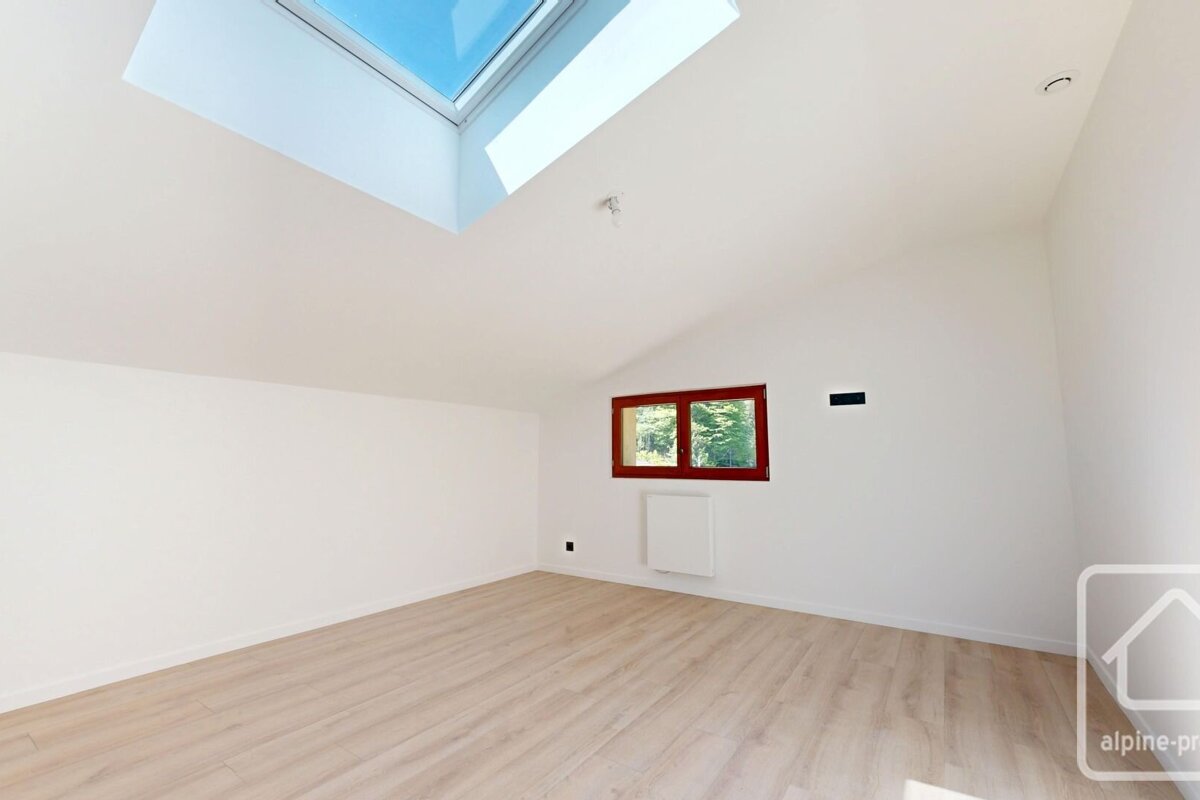 A bright, empty attic room with white walls, a light wood floor, a large skylight, and a red-framed window overlooking trees.