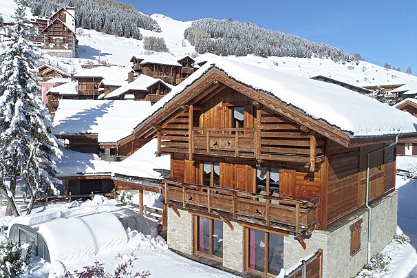 A wooden house with snow on the roof is surrounded by snow covered trees