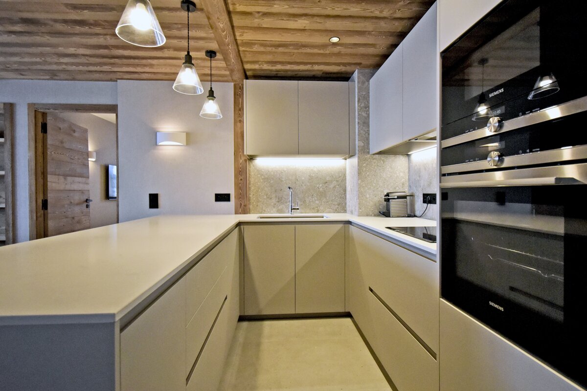 A contemporary U-shaped kitchen with light beige cabinets, built-in ovens, elegant pendant lights, and a rustic wooden ceiling.