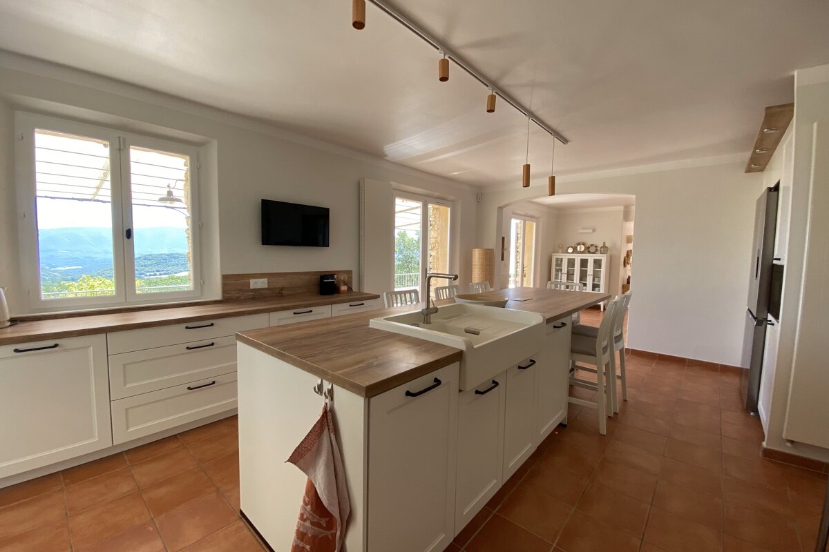 A modern kitchen with white cabinets, wood countertops, and a large island featuring a sink. Enjoy mountain views from the window and a warm terracotta floor.