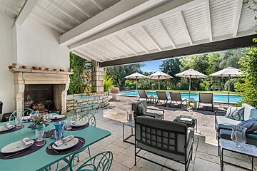 A beautiful covered patio with outdoor dining and lounge furniture, facing a sunny swimming pool area with lounge chairs and umbrellas.