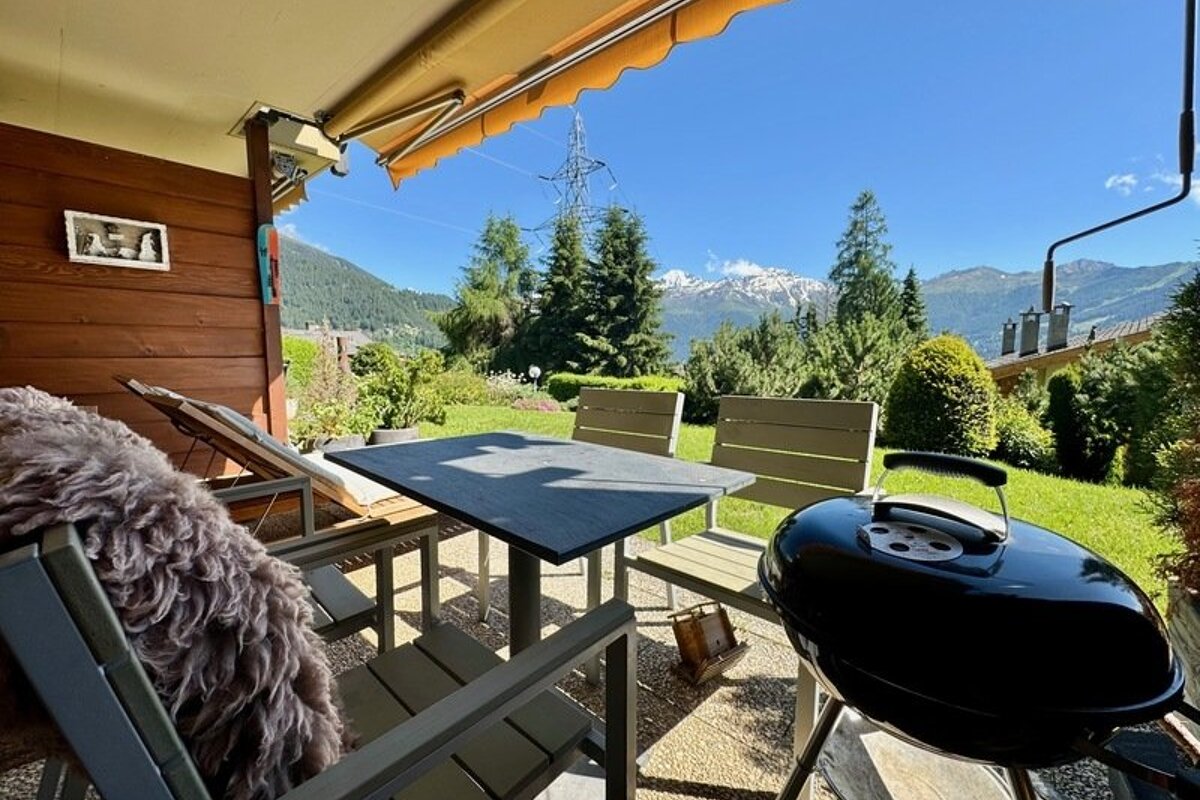 Outdoor patio with a table, chairs, and grill, looking out onto a sunny yard, green trees, and majestic mountains under a clear blue sky.