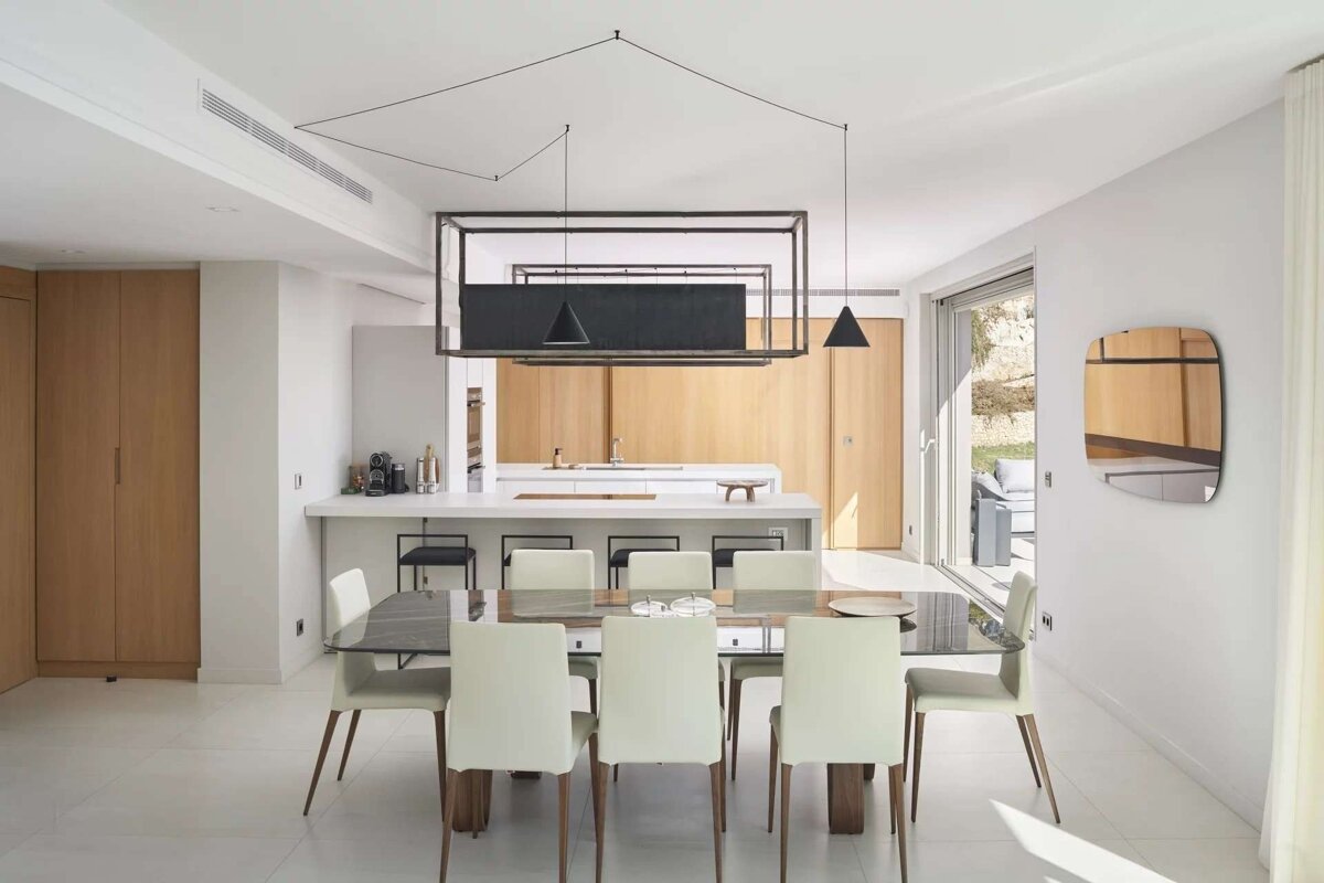 A bright, minimalist open-plan dining and kitchen area with light wood, white surfaces, a large table, and unique black lighting.
