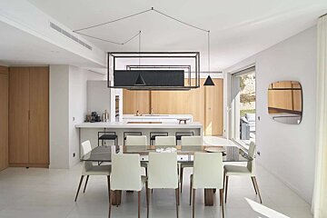 A sleek, modern open-plan dining room and kitchen. It features white walls, light wood cabinets, a striking geometric black chandelier, and leads to an outdoor patio.