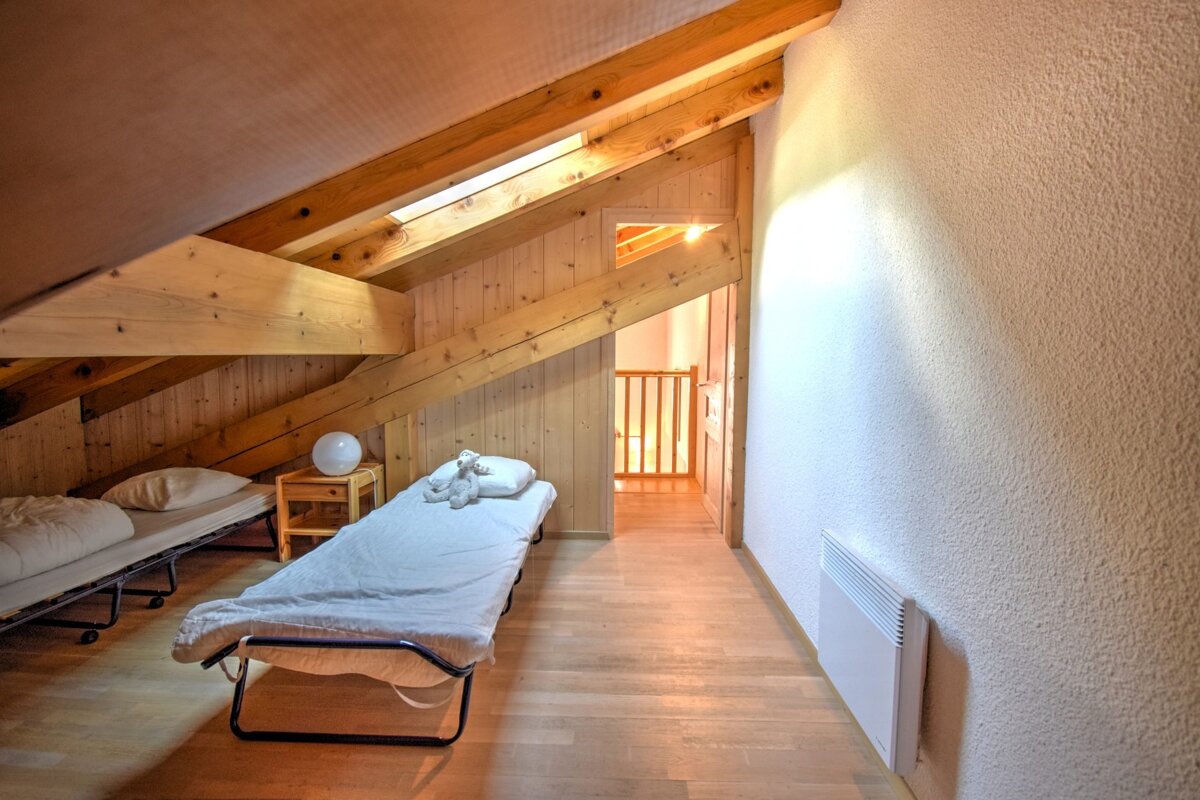 Cozy attic bedroom with exposed wooden beams, two fold-out beds, a nightstand with a lamp, and a hallway entrance. Light wood floors and a white textured wall.