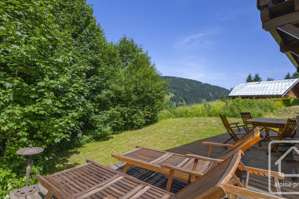 A sunny mountain view from a wooden deck with lounge chairs and a table. Lush green trees and grass frame a distant house under a clear blue sky.