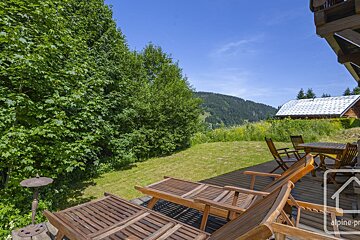 A sunny mountain view from a wooden deck with lounge chairs and a table. Lush green trees and grass frame a distant house under a clear blue sky.