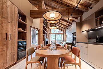 Modern rustic dining room in a chalet with a wooden oval table, colorful chairs, pendant lights, and exposed ceiling beams.