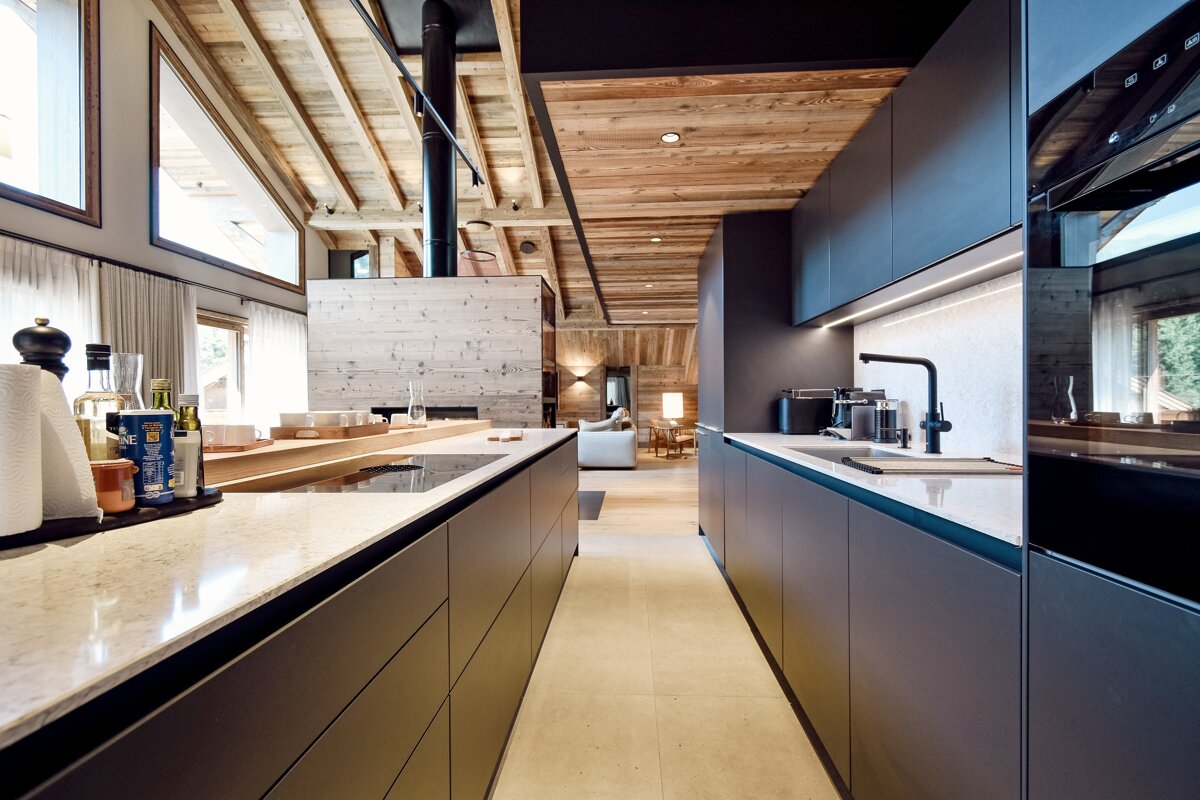 A modern, open-plan kitchen with dark cabinetry, light countertops, and warm wood ceilings, blending contemporary design with rustic chalet style.