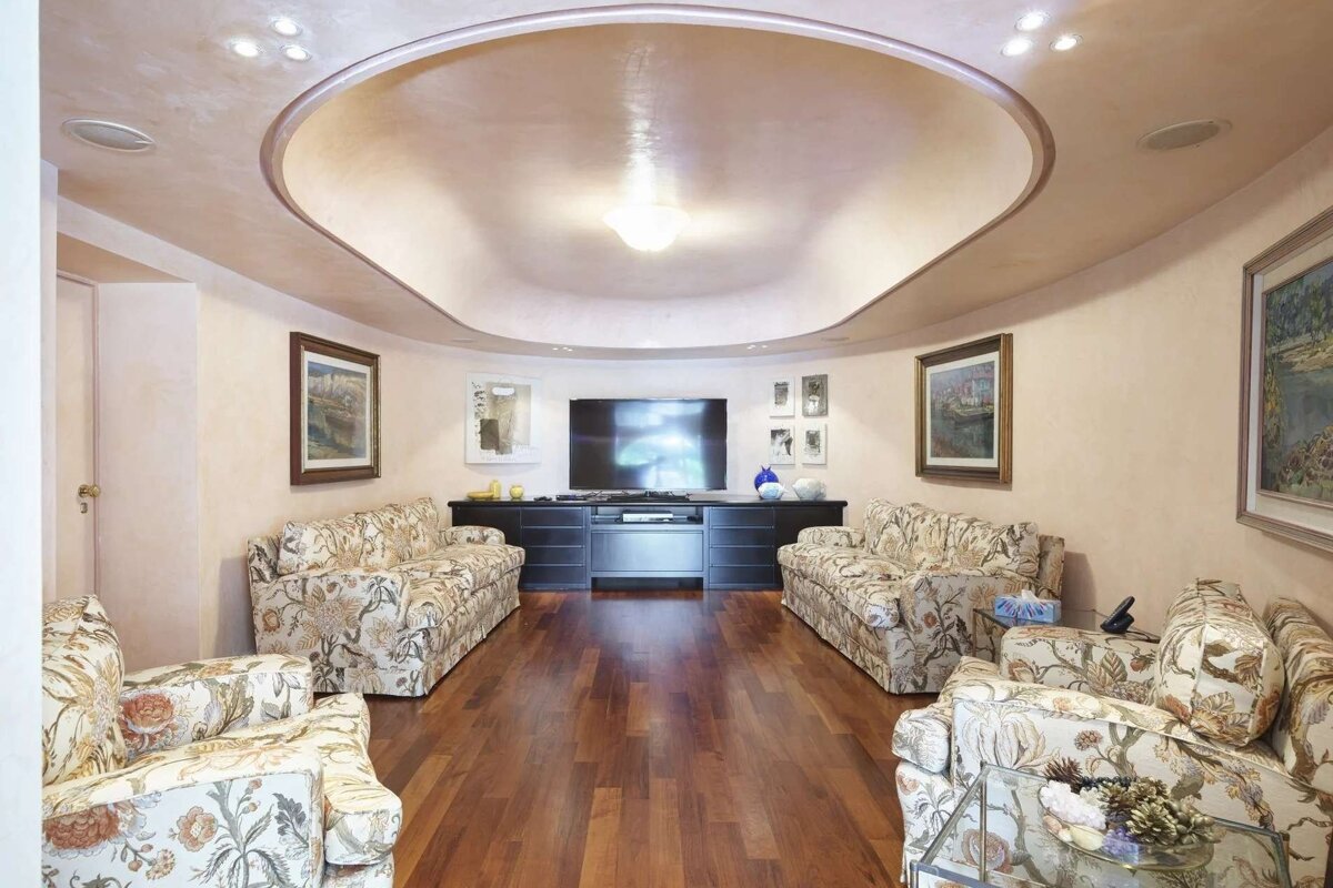 A curved living room with floral sofas facing a large TV. Features hardwood floors, paintings, and a unique cove-lit ceiling design.