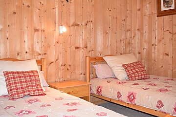 Cozy cabin-style bedroom with knotty pine walls, featuring two twin beds with red plaid pillows and a small wooden nightstand.