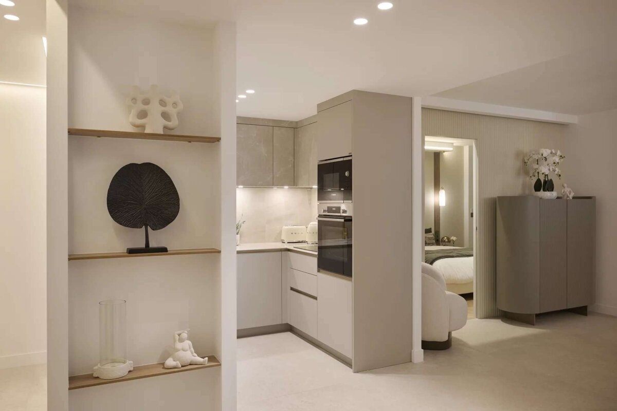 Modern open-plan apartment featuring a light grey kitchen, decorative shelving, and a glimpse of a cozy bedroom.