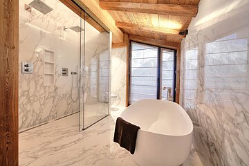 Luxurious bathroom featuring marble walls and floor, a large glass shower, and a modern freestanding tub. Rustic wooden beams and large windows complete the stylish design.