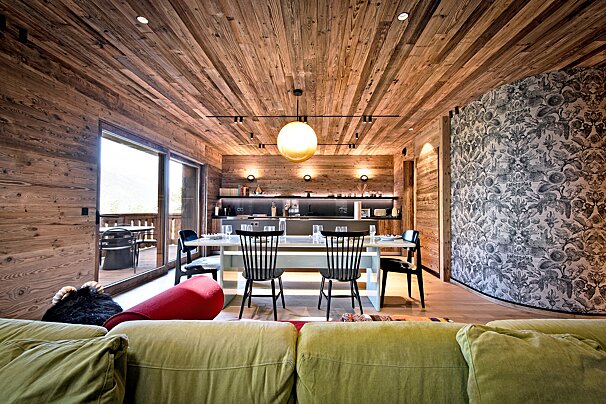 A stylish chalet interior features wood-paneled walls/ceiling, a dining area, modern kitchen, scenic balcony, and unique patterned wallpaper. Cozy green sofa.