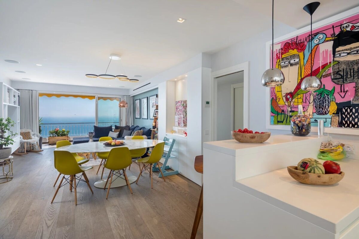 A bright, modern open-plan living/dining room with a sea view balcony, bright yellow chairs, and colorful pop art, featuring hardwood floors.