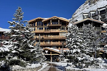 A large wooden ski chalet and neighboring buildings, covered in snow, surrounded by snow-dusted pine trees under a clear blue sky.