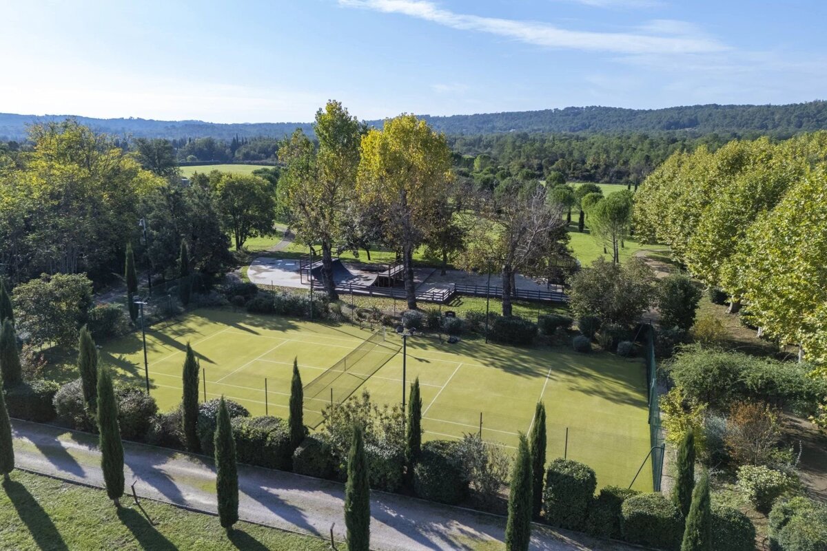 Aerial view of a green tennis court surrounded by tall cypress trees, lush woods, and rolling hills under a blue sky.