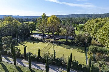 Aerial view of a green tennis court surrounded by tall cypress trees, lush woods, and rolling hills under a blue sky.