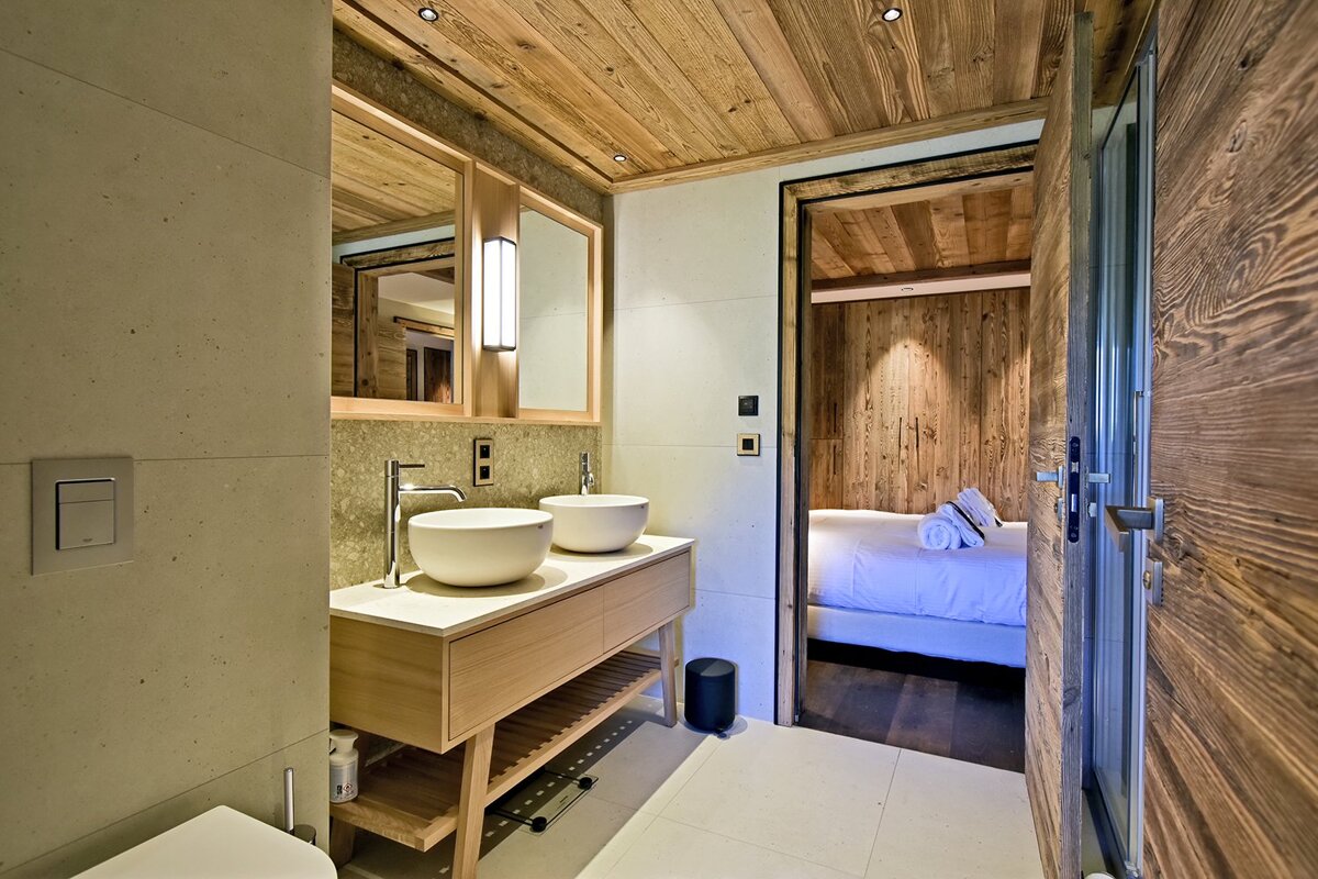 Modern chalet-style bathroom with double vessel sinks, wood ceiling, and a view into a cozy bedroom.