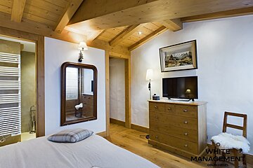 A cozy, rustic-style bedroom featuring a wooden beamed ceiling, white walls, a bed, mirror, TV on a dresser, and a chair with a fur throw.