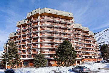 A large, terraced alpine building with numerous wooden balconies sits in a snowy mountain landscape, flanked by pine trees and parked cars under a blue sky.