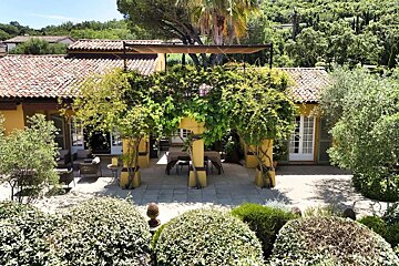 A yellow Mediterranean villa with a red-tiled roof, a vine-covered patio with outdoor dining, and lush green gardens.
