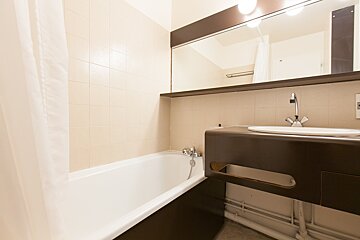 A bathroom featuring a white bathtub with a shower curtain, a dark brown vanity with a white sink, and a large mirror above it. Light-colored tiles line the walls.