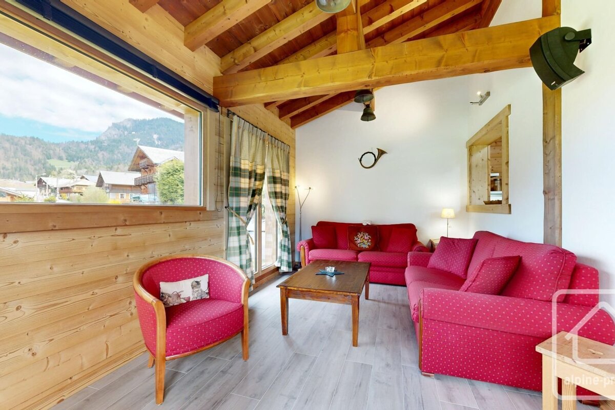 An alpine living room with red seating, exposed wooden beams, and a large window offering a scenic view of a mountain village.