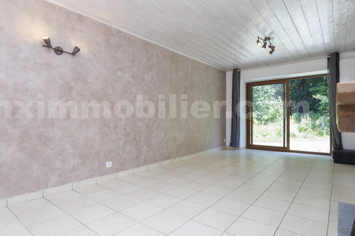 An empty room with light tiled floor, textured beige walls, and wooden slat ceiling. A large glass door reveals a green outdoor view.