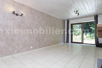 An empty room with light tiled floor, textured beige walls, and wooden slat ceiling. A large glass door reveals a green outdoor view.