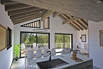 Modern open-plan kitchen, dining, and living area with a high wood-beamed ceiling, large windows overlooking a forest, and a spacious balcony.