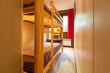 A narrow room features wooden bunk beds with patterned bedding, a window with red curtains, and a small desk, all on a light carpeted floor.
