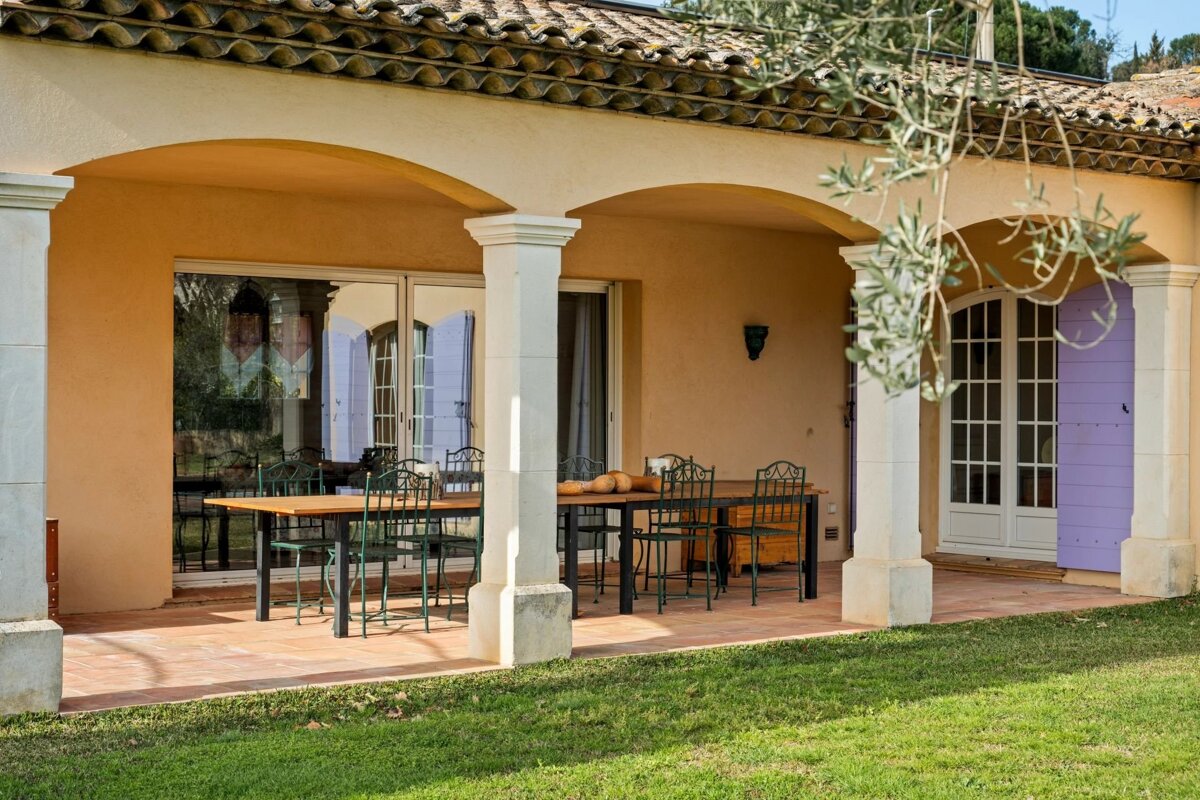 A sunlit covered patio with a long dining table, green chairs, arched openings, and a house with a tiled roof and purple shutters, bordered by green grass.