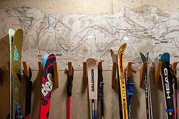 Skis and poles hang on a wall rack beneath a large ski resort map on a concrete wall.