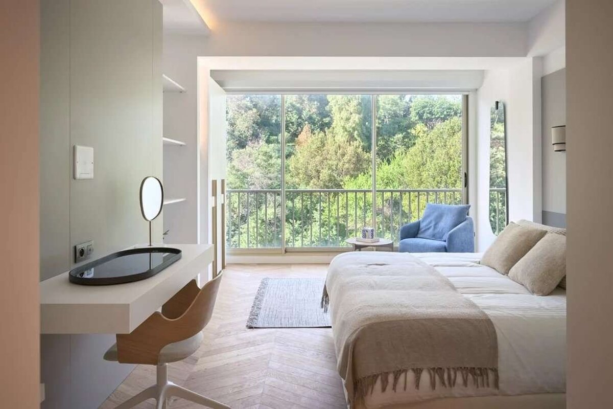 A modern, bright bedroom features a large window overlooking a lush green forest, with a bed, desk, and blue armchair.