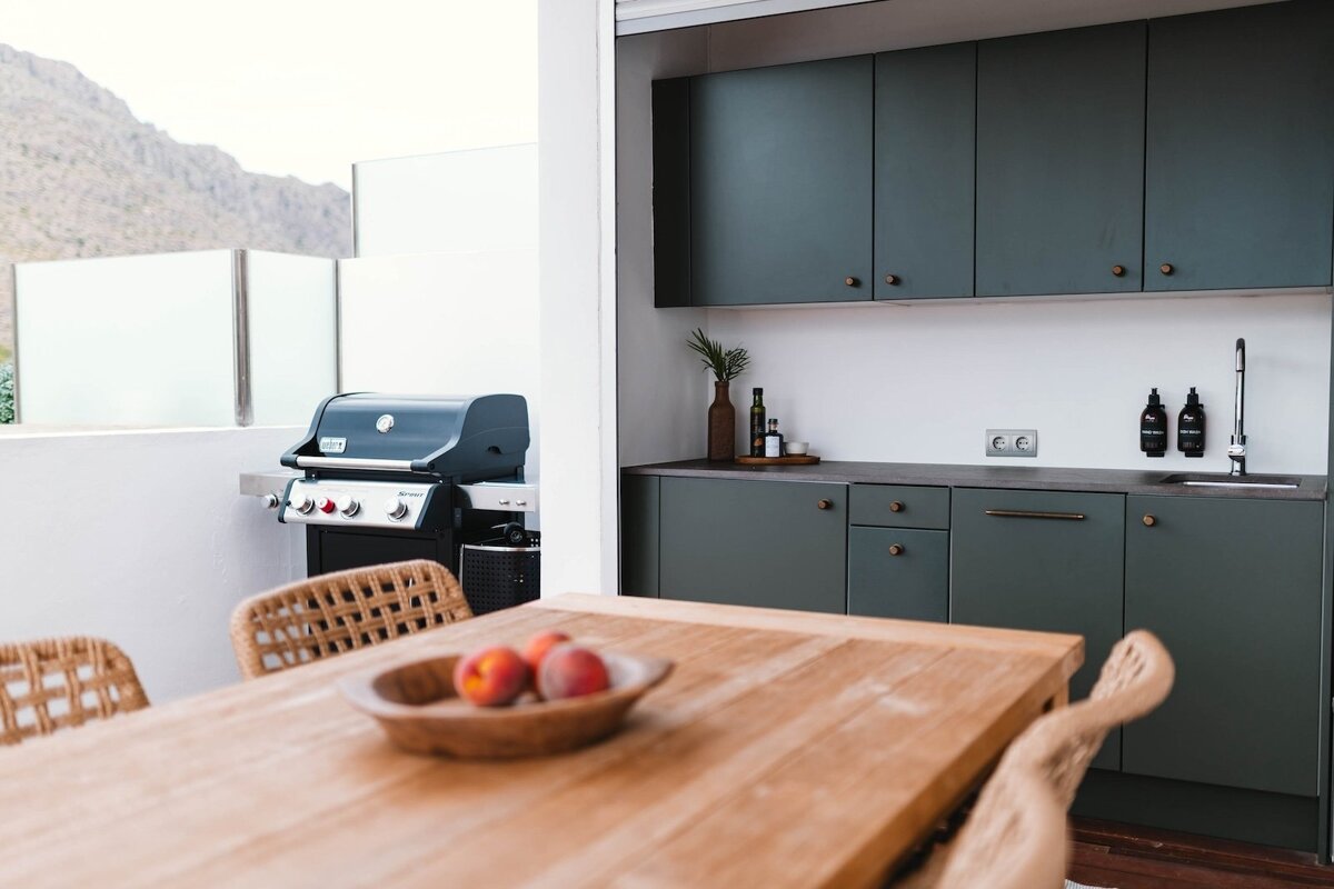 An outdoor kitchen area features a grill, dark green cabinets, and a wooden table with peaches, all set against a distant mountain backdrop.
