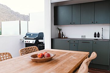 An outdoor dining area with a wooden table, grill, and dark green outdoor kitchen, set against a mountain backdrop.