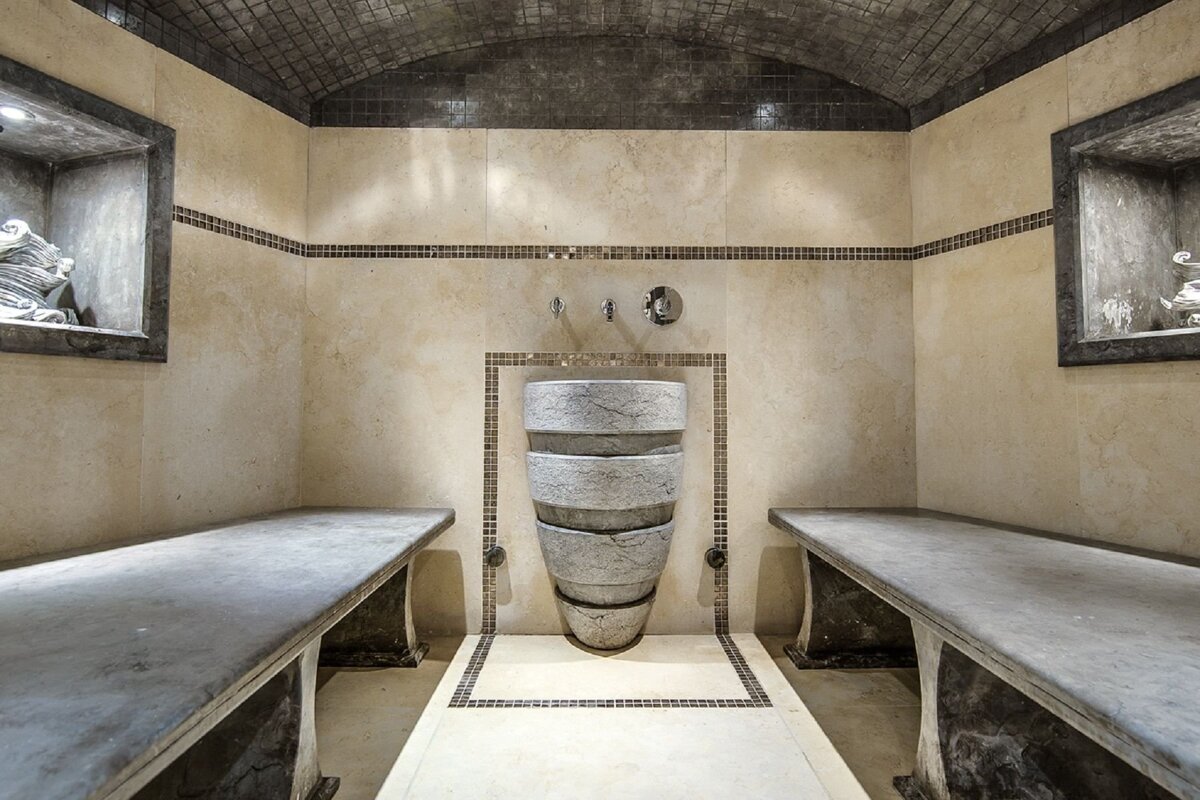 A luxurious hammam with neutral-toned tiled walls, two long stone benches, a unique stacked stone basin, and an arched, mosaic-tiled ceiling.