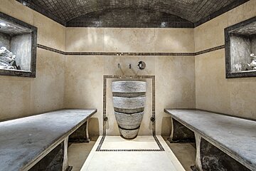 A luxurious hammam with neutral-toned tiled walls, two long stone benches, a unique stacked stone basin, and an arched, mosaic-tiled ceiling.