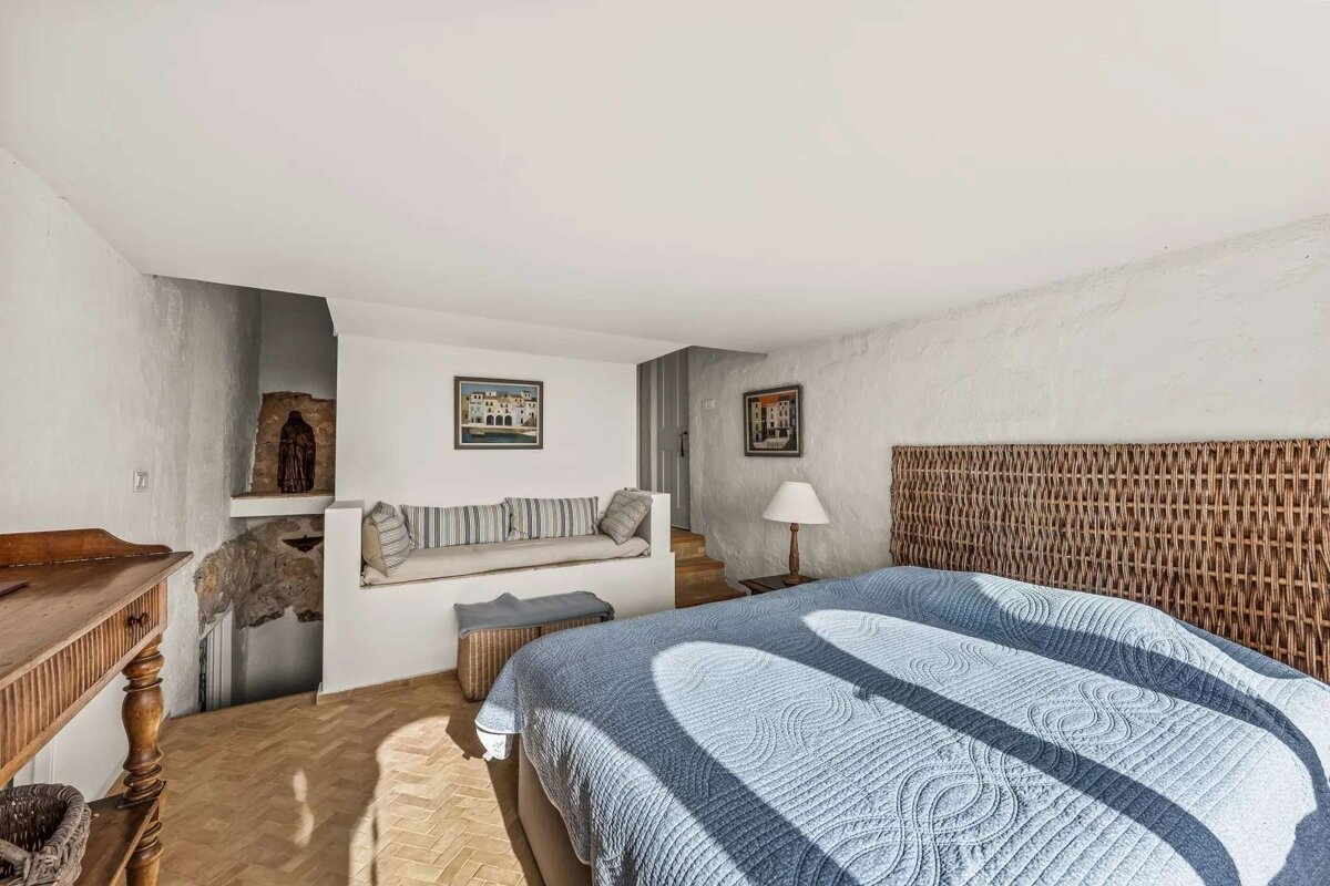 Sunlit bedroom with a blue bed, wicker headboard, white textured walls, a built-in bench, and an exposed stone alcove.