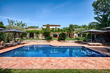 A luxurious villa with a large blue swimming pool, terracotta patio, and lush green garden under a clear sky. Umbrellas and lounge chairs are by the pool.