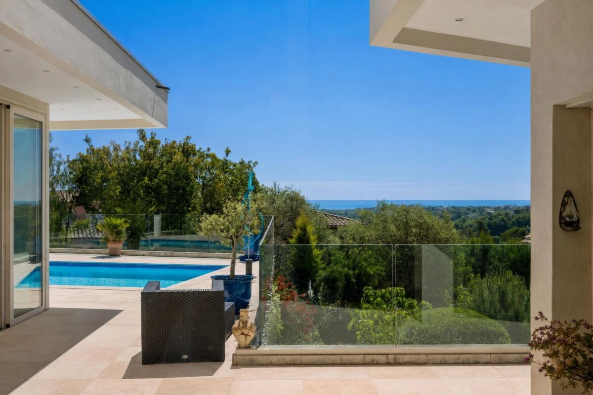 Modern terrace with a pool, glass railing, lush greenery, and a distant sea view under a clear blue sky.