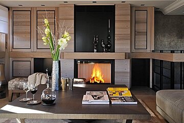 Cozy modern living room with a roaring fireplace, light wood and black walls, a coffee table with wine, flowers, and magazines.