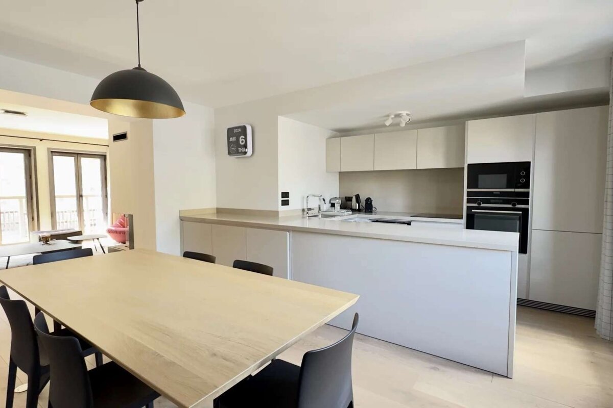 A modern, open-plan kitchen and dining area features a light wood table, sleek white cabinets with built-in appliances, and a black pendant light.